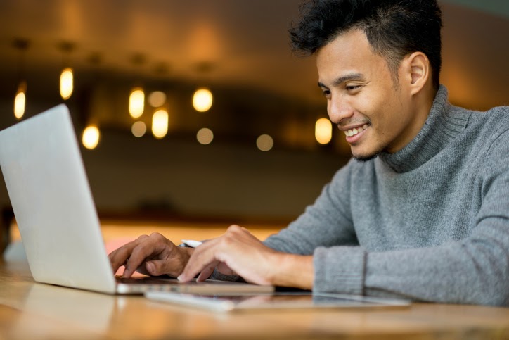A smiling young man typing on his laptop.