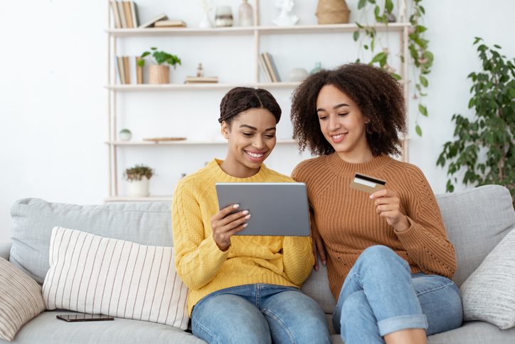 Two smiling students sitting on a couch and holding a credit or debit card and a tablet.