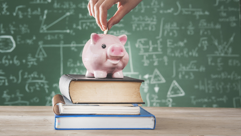 A piggy bank sitting on college textbooks in front of a chalkboard covered with equations.