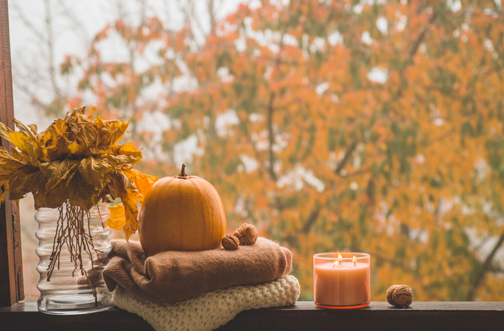 A stack of blankets, a pumpkin, and orange candle on a window sill.