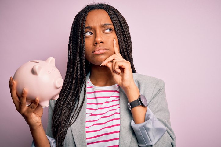 A young woman in business casual attire holding a piggy bank with a quizzical look on her face.